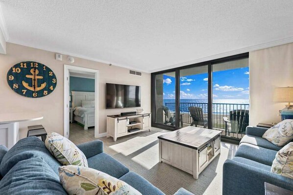 A modern living room with a large TV, comfortable seating, a wooden coffee table, and a balcony with a view. - Sea Colony Ocean 5th floor condo w/ free WiFi, elevator, and balcony (Bethany Beach)