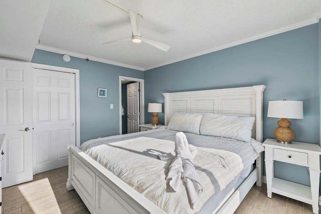 A bedroom with a white bed, two bedside tables with lamps, a ceiling fan, and a door.