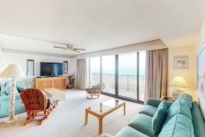 A spacious living room with a sofa, armchair, and coffee table, a television, and a balcony with a view. - Sea Colony oceanfront 8th floor condo with shared sauna, tennis court, & pool (Bethany Beach)