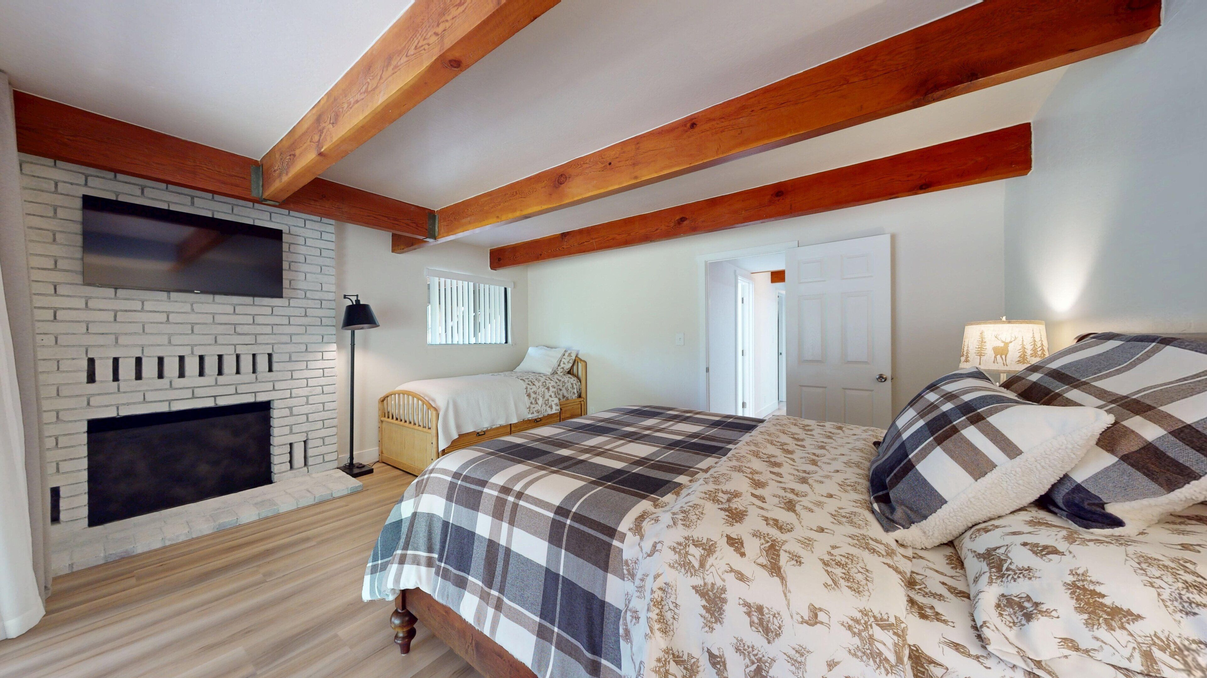Een slaapkamer met een open haard, een bed met ruitdessin beddengoed, houten balken en een witte bakstenen muur.