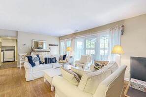 Een woonkamer met een bank, fauteuils, een salontafel en een televisie. Er is een keuken met een koelkast en een eettafel.