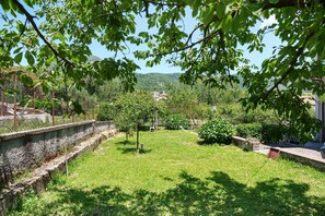 A grassy outdoor area with trees, a stone wall, and a view of distant buildings. - Olympus Apartment – Agerola (Pianillo)