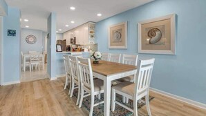 Een eetgedeelte met een houten tafel en witte stoelen, een keuken op de achtergrond en kunstwerken in lijsten aan de muren.
