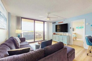 Ein Wohnzimmer mit einer Sofagarnitur, einem Fernseher, einem Deckenventilator und einer Glastür zum Außenbereich.