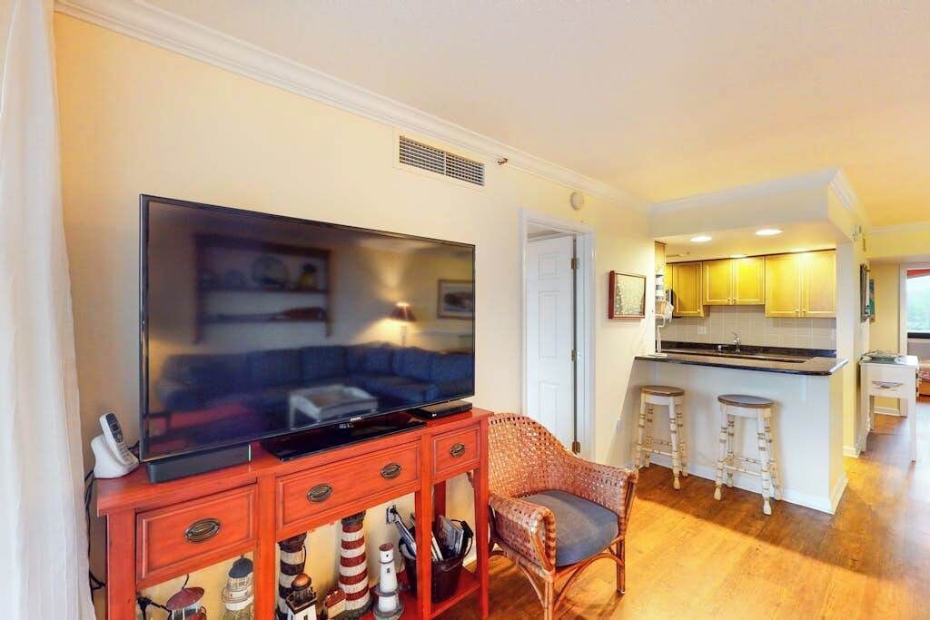 Un salon équipé d’un téléviseur à écran plat, d’un fauteuil en osier et d’un coin cuisine avec des meubles en bois et un tabouret de bar.