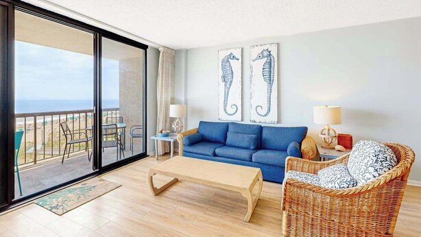 Een kamer met balkon, een blauwe bank, een rieten stoel en twee schilderijen van paard van zee.