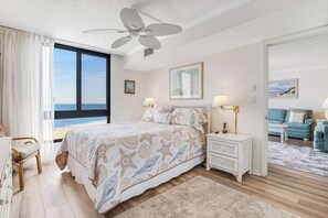 Un dormitorio con cama, mesitas de noche, ventilador de techo, vista al mar y una sala de estar con sofá.