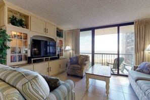 Ein Wohnzimmer mit Fernseher, Sofa, Sessel und Tisch. Ein Balkon mit Aussicht ist vorhanden.