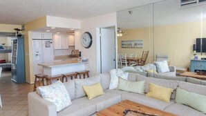 A living room with a sofa, coffee table, and kitchen area with a refrigerator and clock. - Sea Colony oceanfront, 7th-floor condo w/ pool and ocean view (Bethany Beach)
