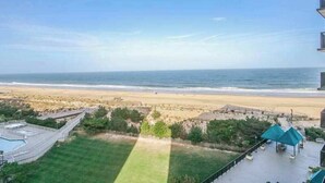 A view of a beach with a clear sky, a pool area, and a building with a balcony overlooking the ocean. - Sea Colony oceanfront, 7th-floor condo w/ pool and ocean view (Bethany Beach)