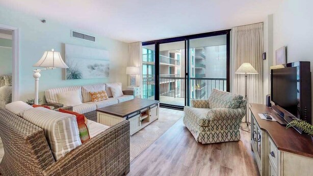 A modern living room with a sofa, armchair, and coffee table, a television, and a large window with a view of buildings. - Sea Colony Ocean 6th floor condo w/ gym, pool, and tennis court (Bethany Beach)