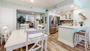 A modern kitchen with white cabinetry, a central island, and a dining area with white chairs. - Sea Colony Ocean 6th floor condo w/ gym, pool, and tennis court (Bethany Beach)