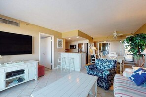 A living room with a TV, white entertainment unit, and a sofa. There is a kitchen area with a bar stool and a dining table with chairs. A ceiling fan is visible. - Sea Colony Ocean 3rd floor condo w/ tennis court, elevator, and balcony (Bethany Beach)