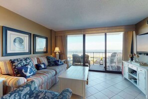 Sala de estar com sofá, mesa de centro e televisão. Quadros emoldurados na parede, varanda com cadeiras e vista para o mar.