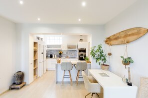 A modern kitchen with white cabinetry, a dining area with two chairs, a surfboard decoration, and a potted plant. - Villa Cannes 10 min from center - Jacuzzi & Sauna (Le Cannet)
