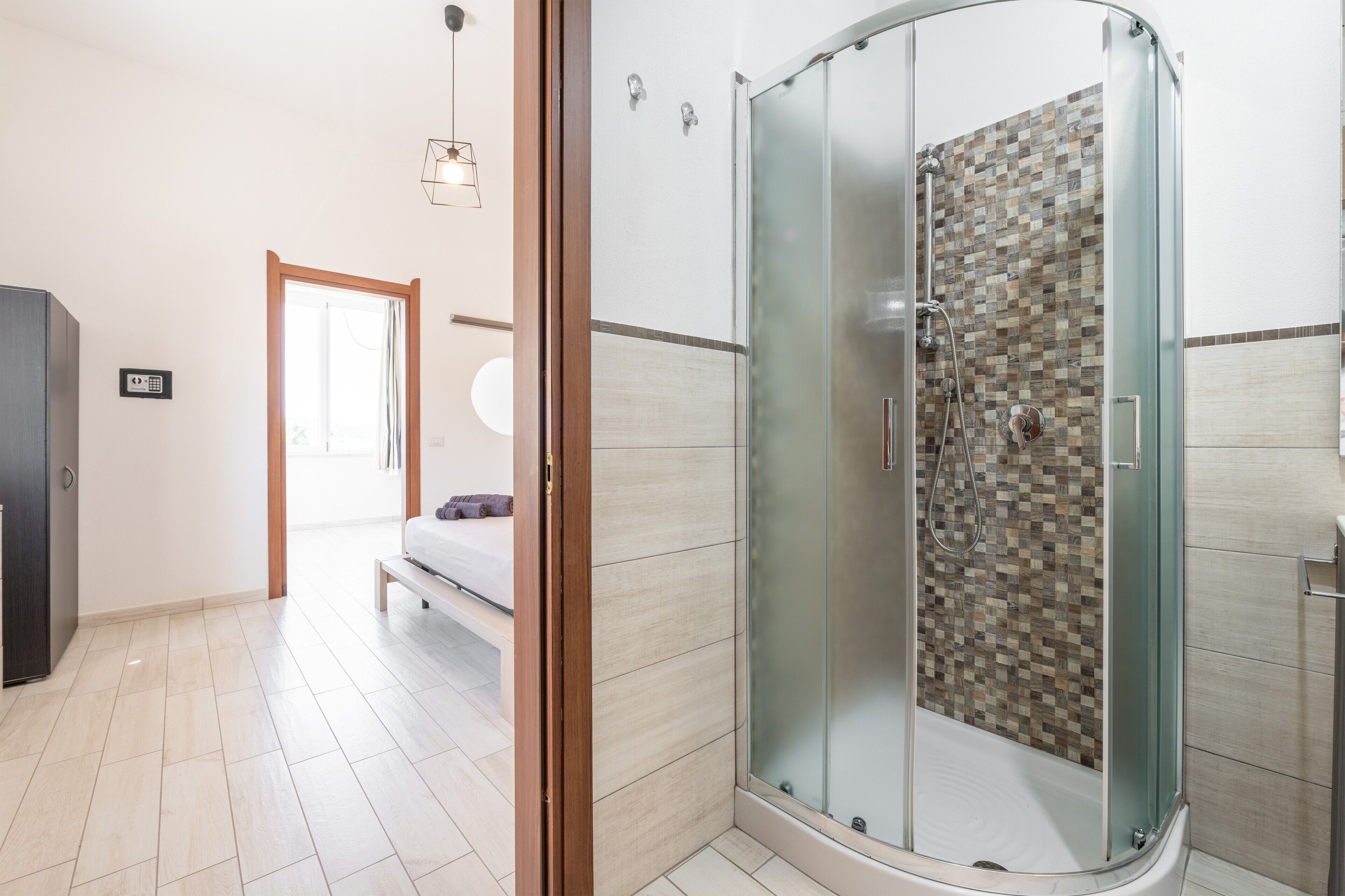 A modern bathroom with a glass-enclosed shower, a visible toilet, and a bed in the adjacent room.