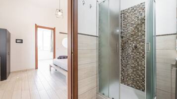 A modern bathroom with a glass-enclosed shower, a visible toilet, and a bed in the adjacent room.
