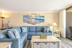 A living room with a blue sofa, a coffee table, wicker baskets, a wall-mounted painting, a lamp, and a small table with a decorative item. - Sea Colony Ocean 1st-floor condo with elevator, pool, & basketball court (Bethany Beach)