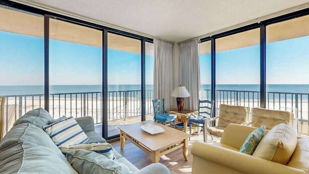 A room with a large window overlooking a beach, a sofa, a table, and two chairs. - Sea Colony 5th Floor Ocean View Condo with Gym (Bethany Beach)