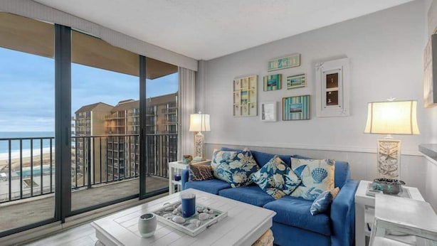 A modern living room with a blue sofa, a white coffee table, and a balcony with a view of the beach and buildings. - Sea Colony 7th-floor ocean-view condo with elevator & gym (Bethany Beach)