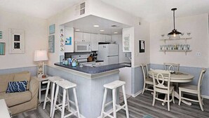 A compact kitchen with white cabinets, a central island with stools, and a dining area with a round table and chairs. - Sea Colony 7th-floor ocean-view condo with elevator & gym (Bethany Beach)