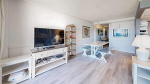 Un soggiorno moderno con una TV a schermo piatto, un tavolino consolle bianco, una mensola in vimini, un tavolo da pranzo bianco e una lampada.