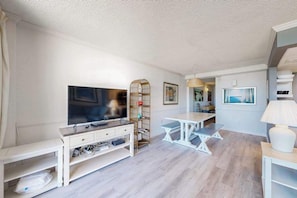 Ein modernes Wohnzimmer mit Flachbildfernseher, weißem Konsolentisch, Weidenregal, weißem Esstisch und einer Lampe.