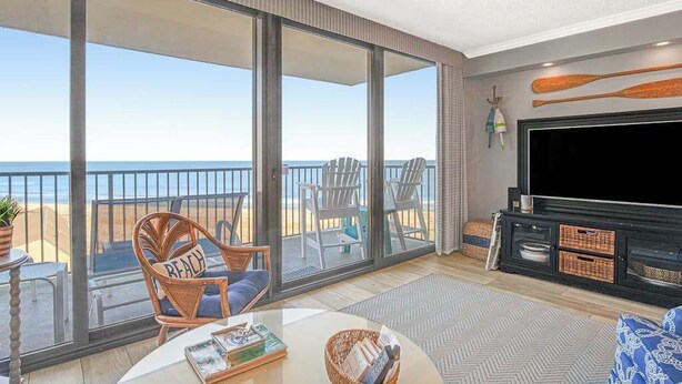 A spacious living room with a large TV, a balcony with chairs, and a view of the beach. - Sea Colony Ocean 7th-floor condo with a gym & a tennis court (Bethany Beach)