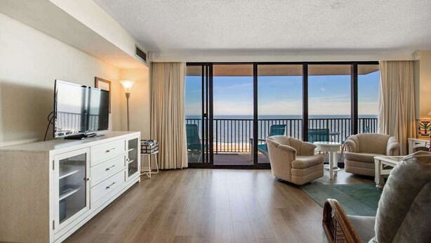Quarto de hotel com varanda, televisão, sofá e mesa de jantar.