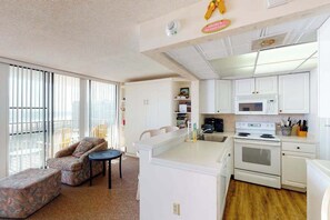 A compact living space with a kitchenette, a sofa, a small table, and a view through large windows. - Sea Colony 7th floor condo w/ shared basketball court, elevator, and ocean view (Bethany Beach)