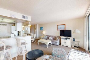 A living room with a sofa, chairs, a coffee table, a television, and a dining area with a table and chairs. - Sea Colony 7th floor condo w/ shared basketball court, elevator, and ocean view (Bethany Beach)