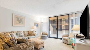 Un salon comprenant un canapé, un pouf, des fauteuils, un téléviseur et une porte-fenêtre coulissante donnant sur un balcon.