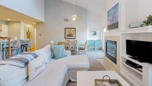 A modern living room with a sofa, TV, ceiling fan, dining area, and artwork. - Sea Colony Tennis 2nd-floor condo with fireplace, basketball court & sauna (Bethany Beach)