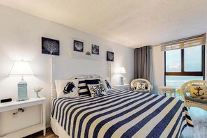 A hotel room with a bed, two bedside tables, a chair, a window with a view, and wall art. - Sea Colony 5th floor condo w/ tennis court, elevator, and gym - ocean view (Bethany Beach)