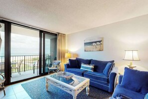 A living room with a blue sofa, a glass coffee table, and a view of the beach through a sliding door. - Sea Colony 5th floor condo w/ tennis court, elevator, and gym - ocean view (Bethany Beach)