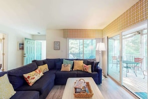 A living room with a dark blue sectional sofa, a coffee table with a basket, and a large window with a view of the outdoors. - Sea Colony Tennis First-floor condo with pool, hot tub & central AC (Bethany Beach)