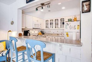 A kitchen with white cabinets, a microwave, a refrigerator, and a bar area with blue chairs. - Sea Colony Tennis townhouse with basketball court, pool & sauna (Bethany Beach)