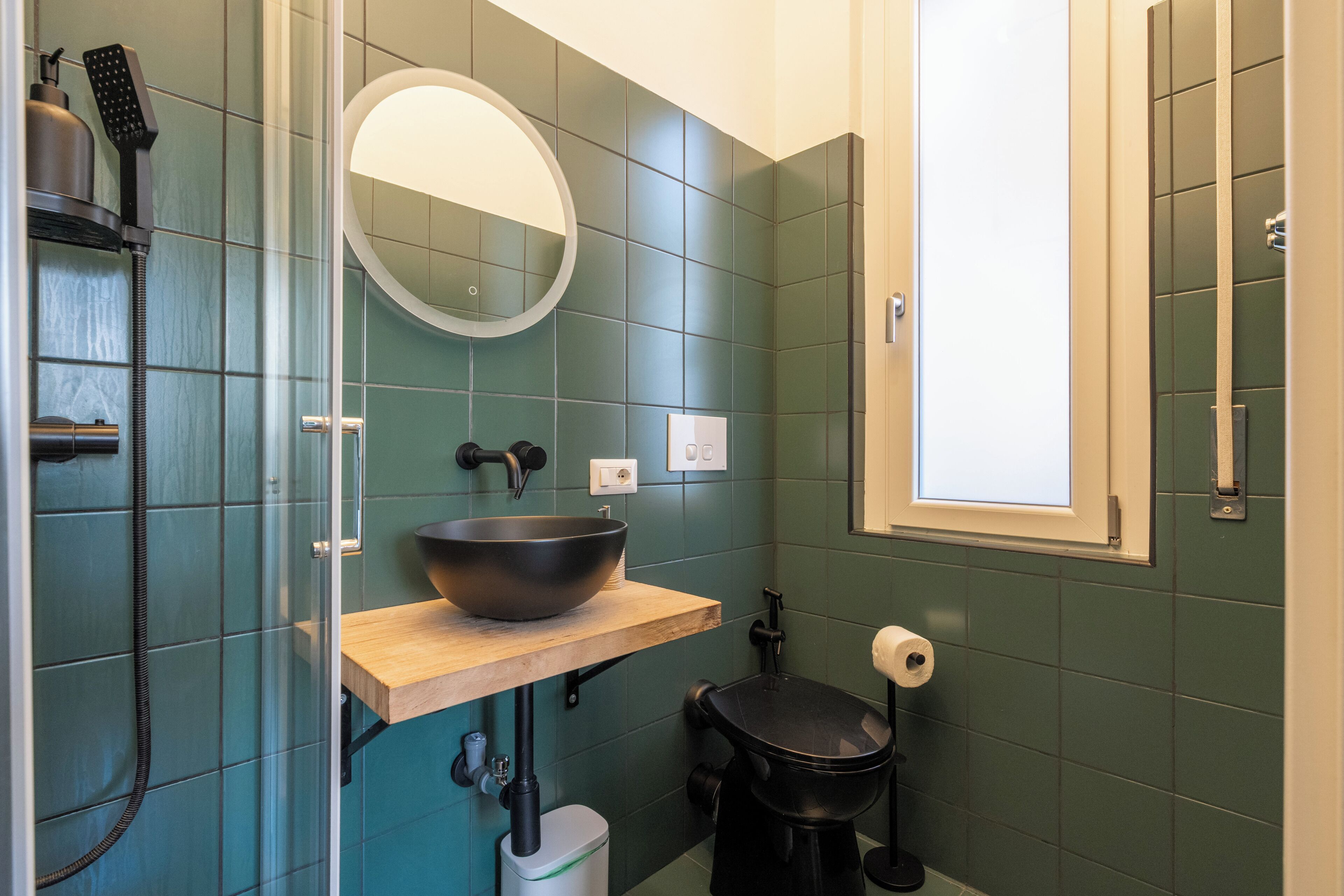 Un baño con ducha envuelta en cristal, tocador de madera con lavabo, inodoro negro y espejo sobre el lavabo.
