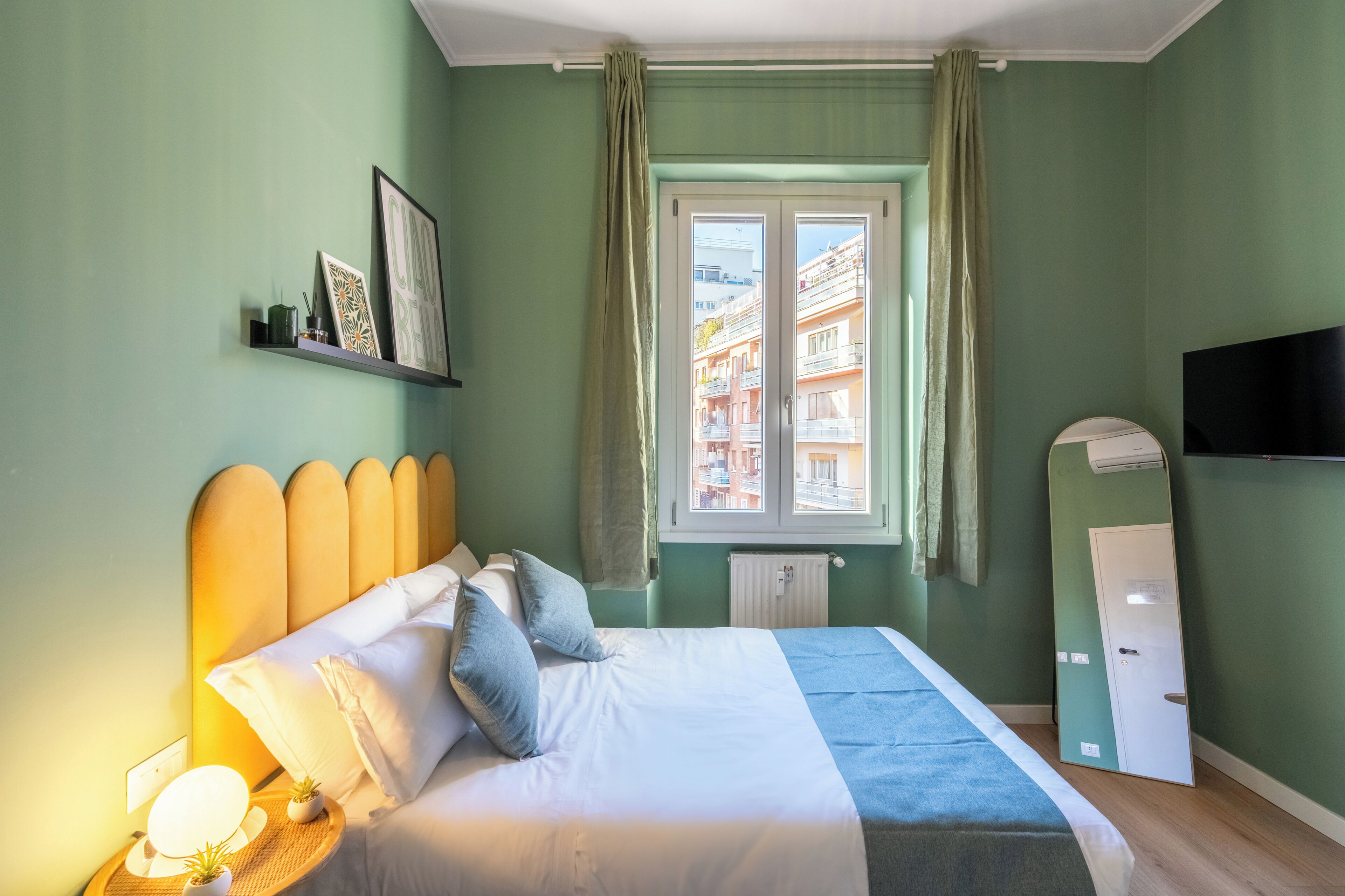 Une chambre à coucher avec un lit, une table de chevet, une lampe, une fenêtre et un téléviseur fixé au mur.