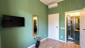 A room with a flat-screen TV, a mirror, a small table, a door leading to a bathroom, and a shower area. - ReRent Rooms 169 Medaglie D'oro (Rome)