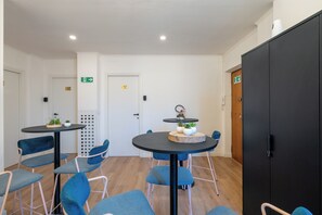 Quarto com duas mesas redondas, cadeiras azuis, um guarda-roupa preto e uma porta branca com sinal de saída.