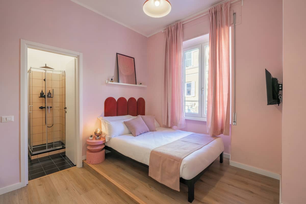 A bedroom with a bed, a shower, a window with curtains, and a mounted TV.