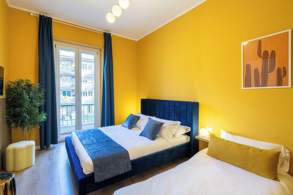 A hotel room with a bed, a bedside table, a nightstand, a potted plant, and a window with curtains.