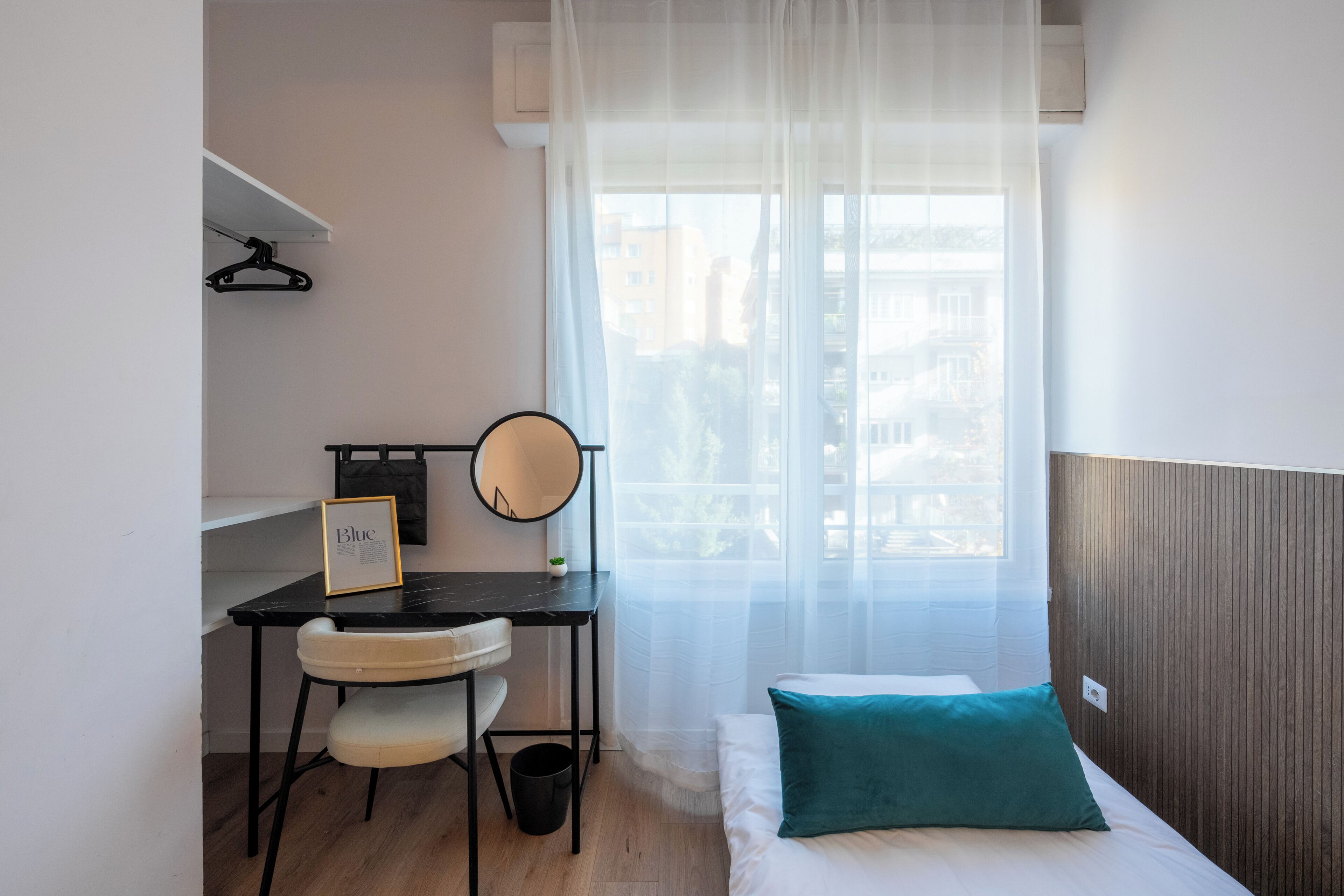 A bedroom with a bed, a desk with a chair, a bedside table, a mirror, and a window with sheer curtains.