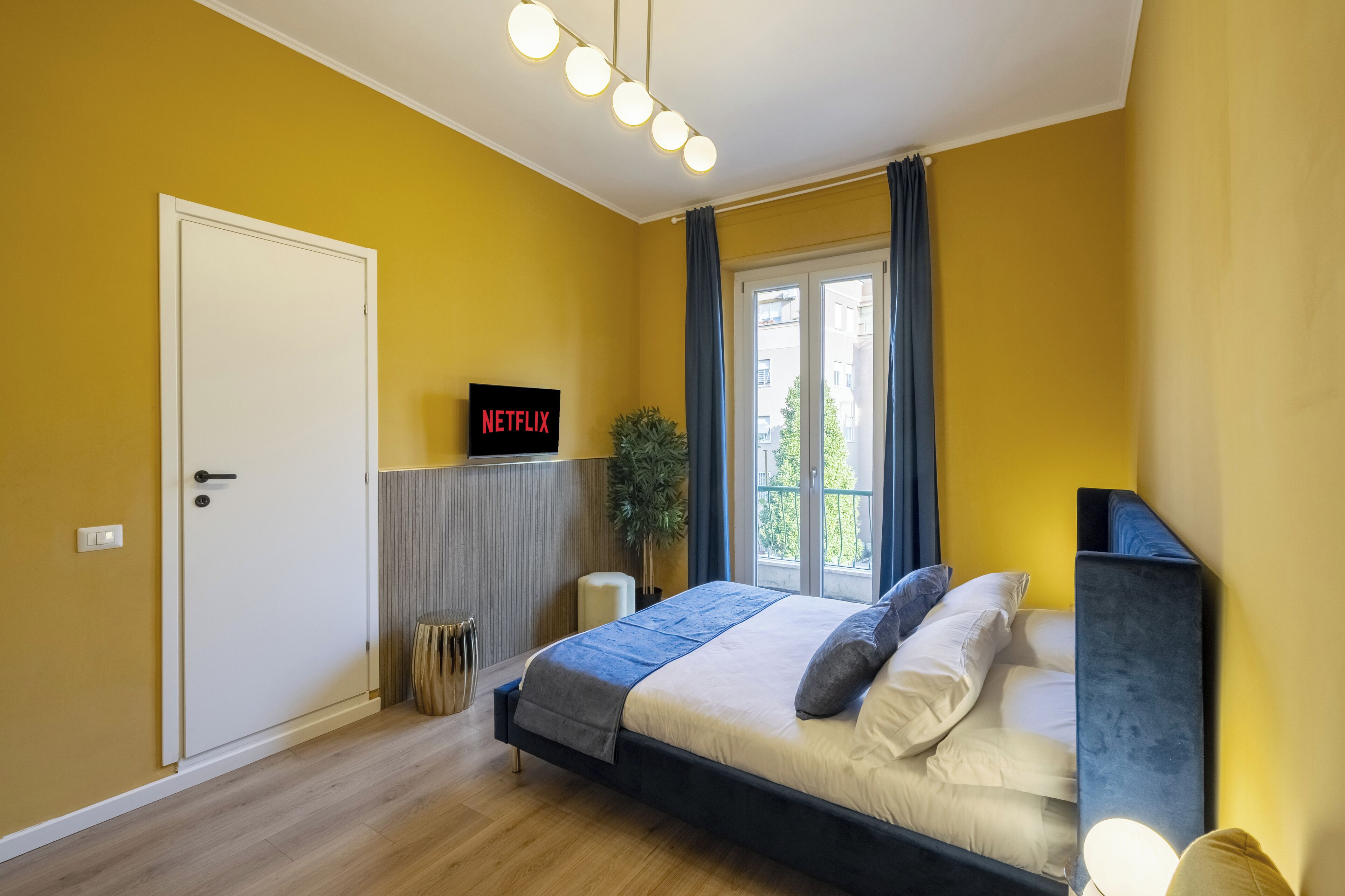 A bedroom with a bed, a wall-mounted Netflix sign, a potted plant, and a window with curtains.
