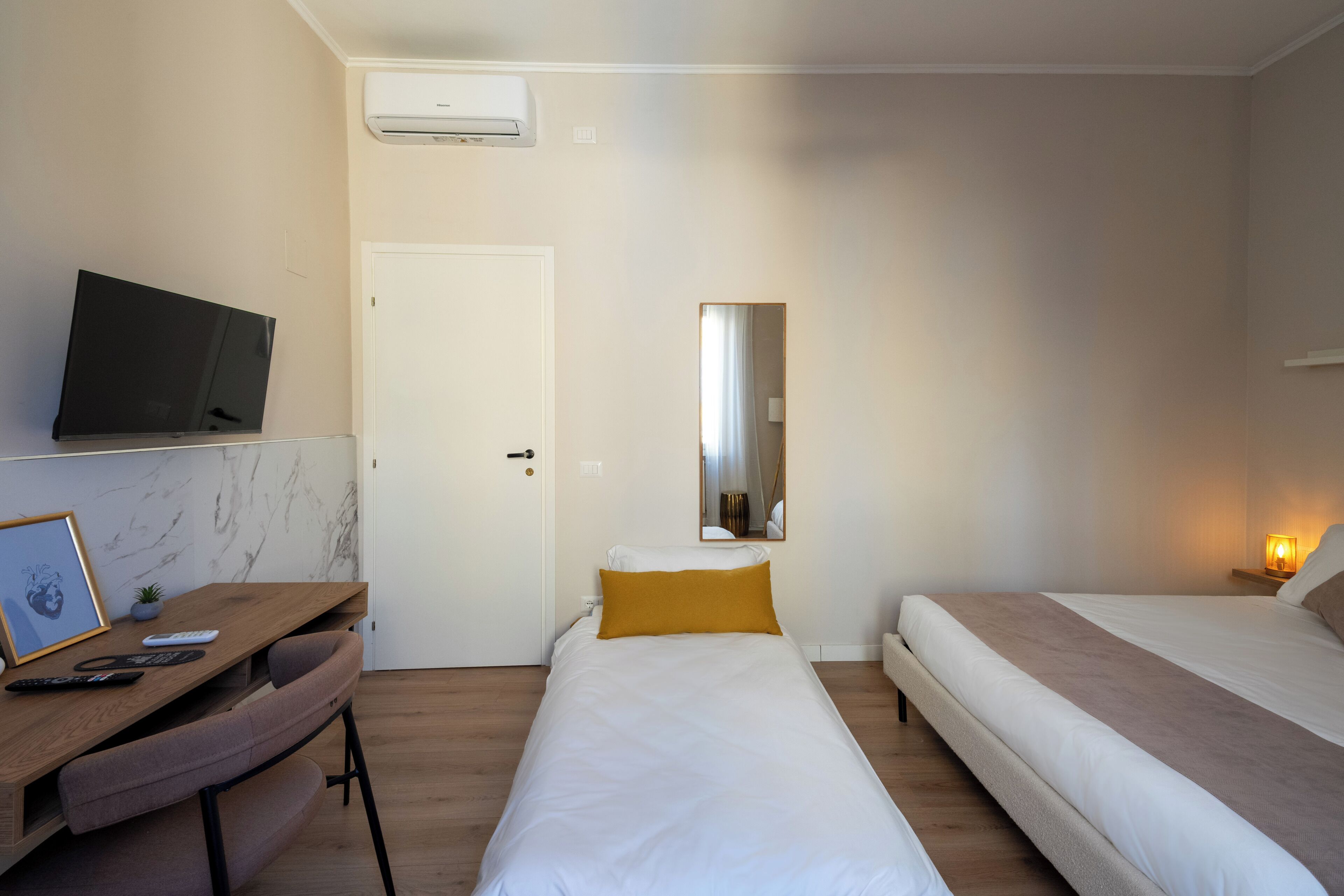 A hotel room with a bed, bedside table, TV, desk with chair, and a framed picture.