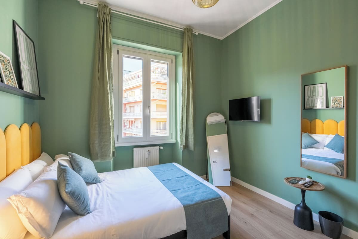 A bedroom with a bed, a window with curtains, a television, a mirror, and a small table with a plant.