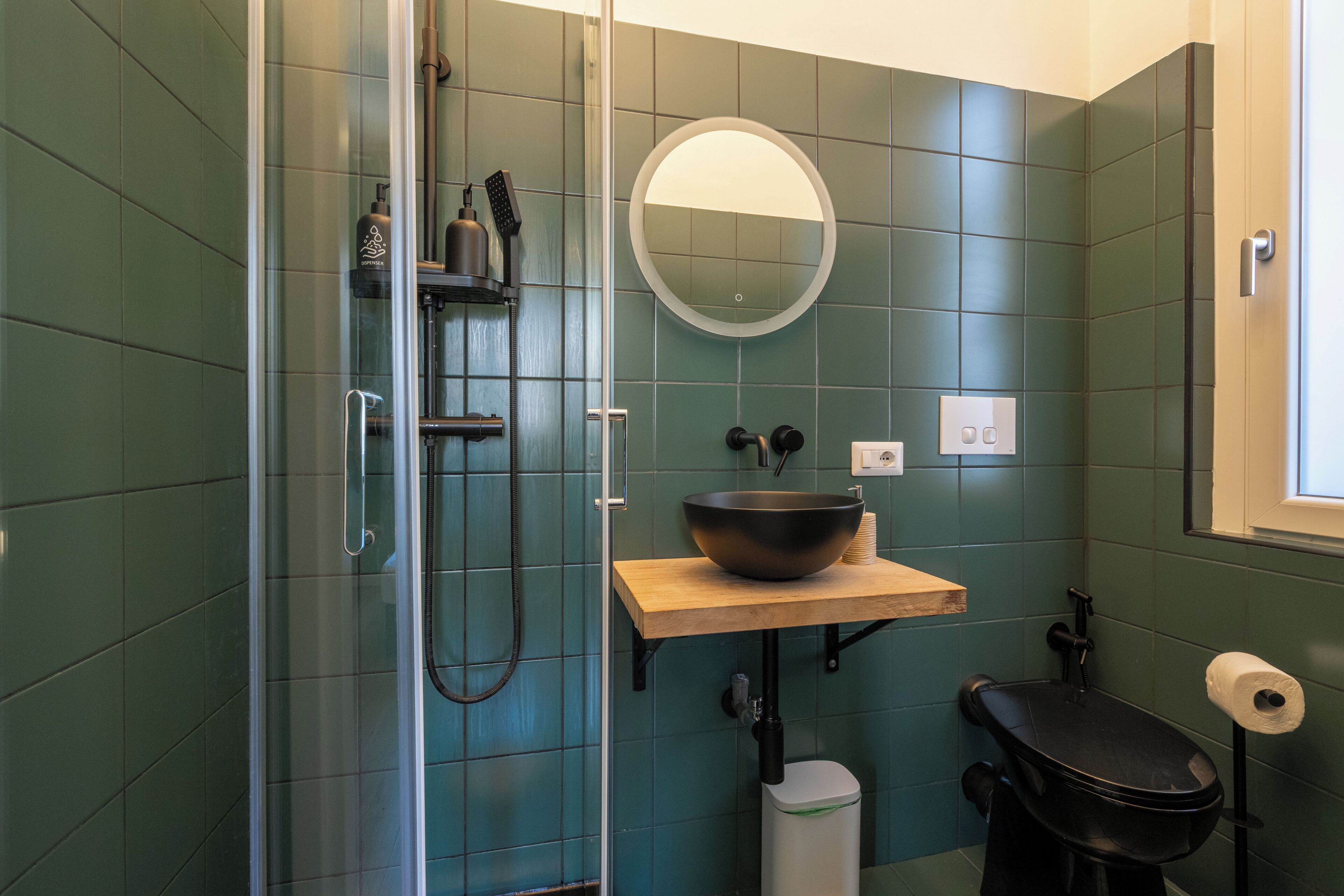A bathroom with a glass shower enclosure, a wooden sink with a black basin, a round mirror, and a toilet with a black seat.