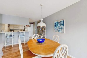 A dining area with a wooden table, white chairs, a blue bowl, a white pendant light, and a kitchen in the background. - Sea Colony Tennis 1st-floor condo sauna & pool (Bethany Beach)