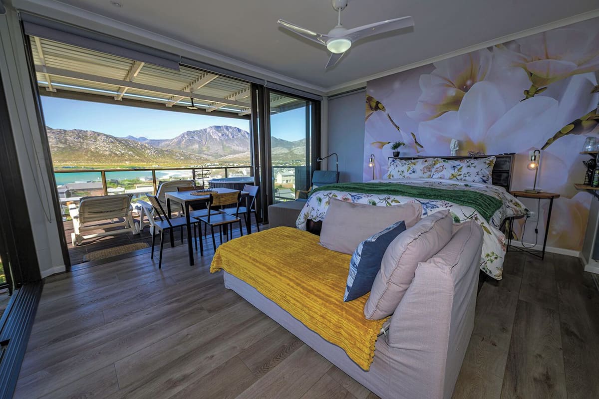 A bedroom with a bed, a desk, and a chair, a balcony with a view of mountains and water, and a ceiling fan.